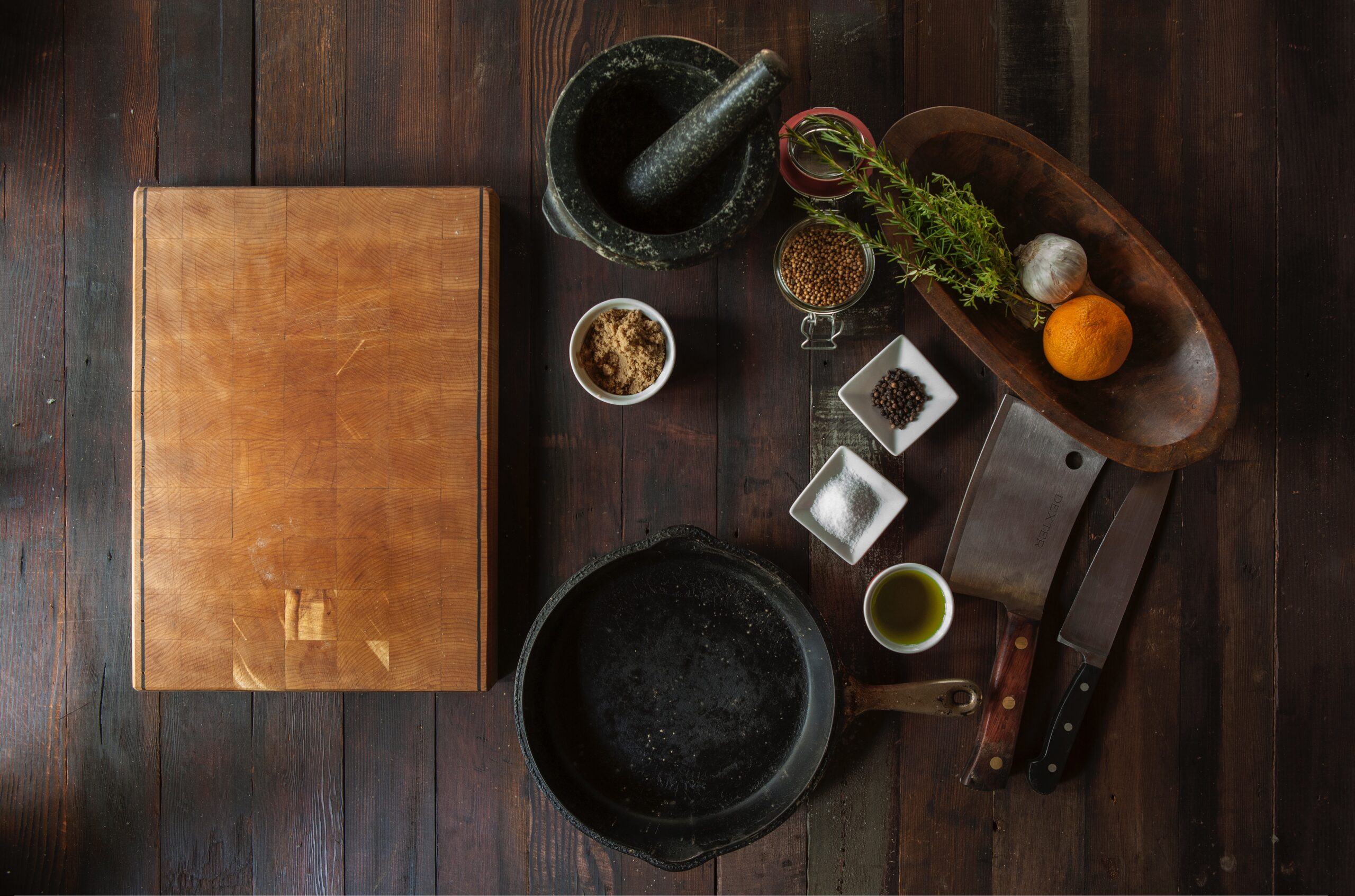 Plano cenital de una mesa de madera con ingredientes para una receta de setas con queso de cabra y jamón ibérico: cuchillos, cuenco con cebolla y romero, mortero, especias, y una sartén