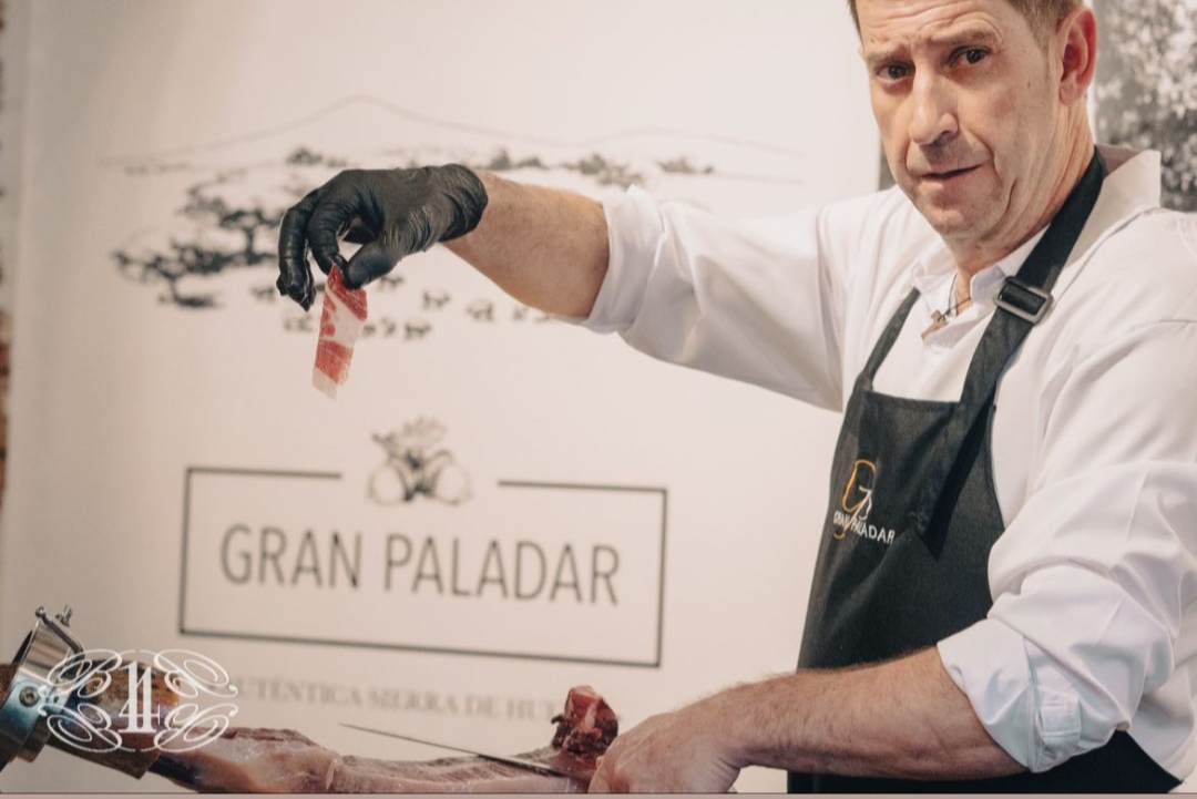 Persona sosteniendo una loncha de jamón con delantal de Gran Paladar, y la palabra 'EVENTOS' escrita en el centro, sobre un fondo difuminado con el logo de Gran Paladar